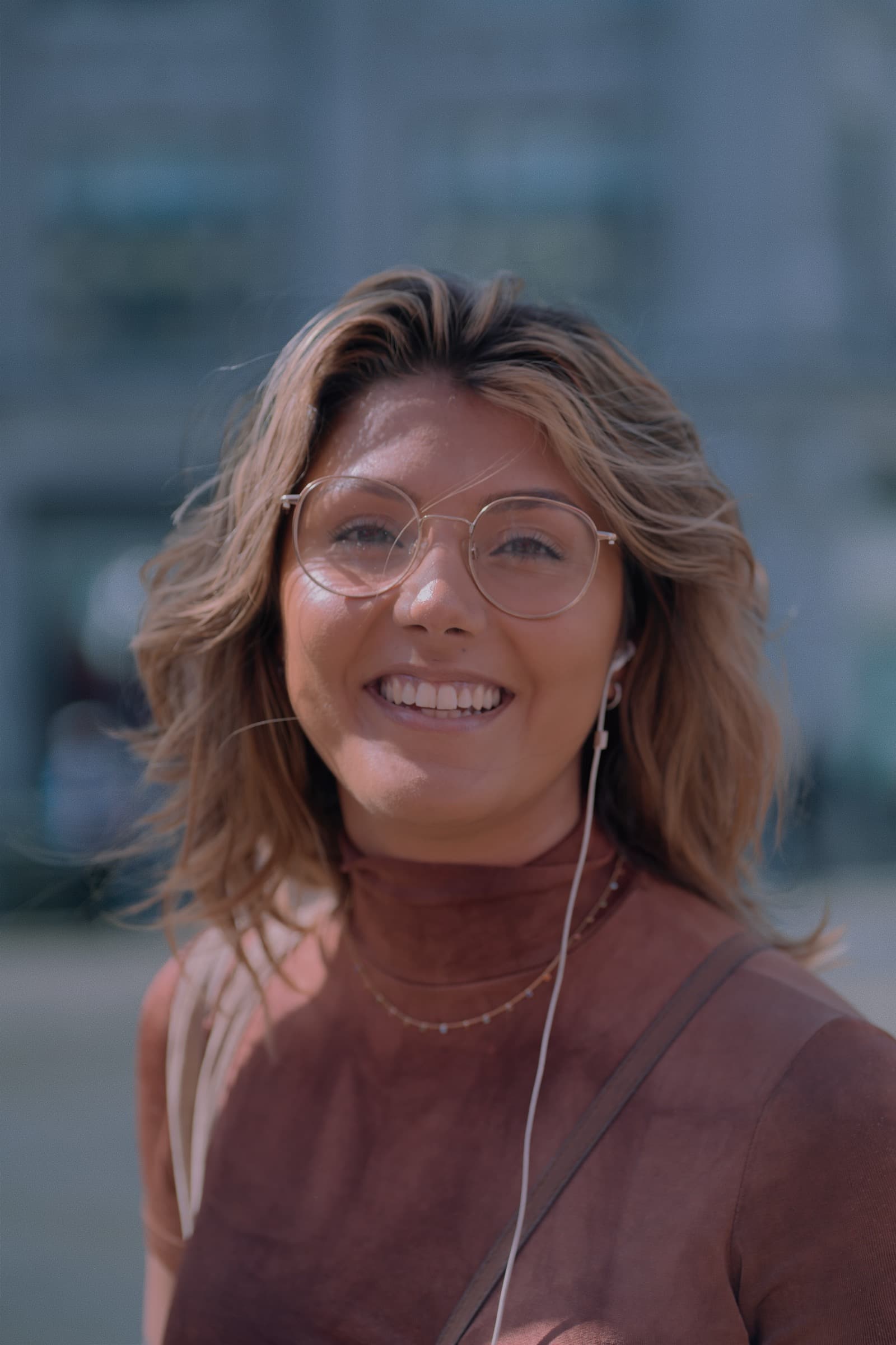 Young woman with round glasses and earphones smiling softly on a city street