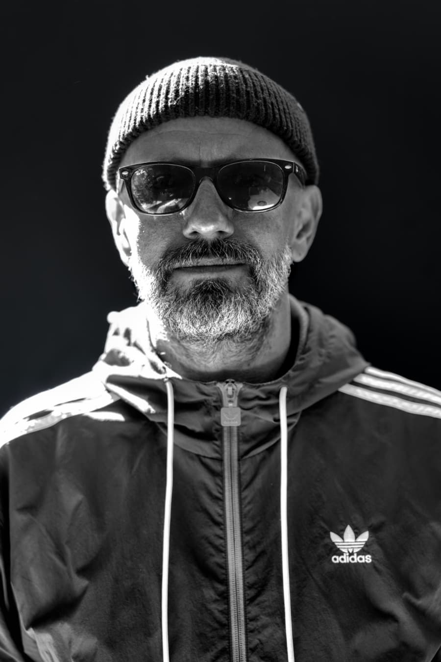 Black and white close-up portrait of a man in a beanie and Adidas jacket with a calm, composed gaze
